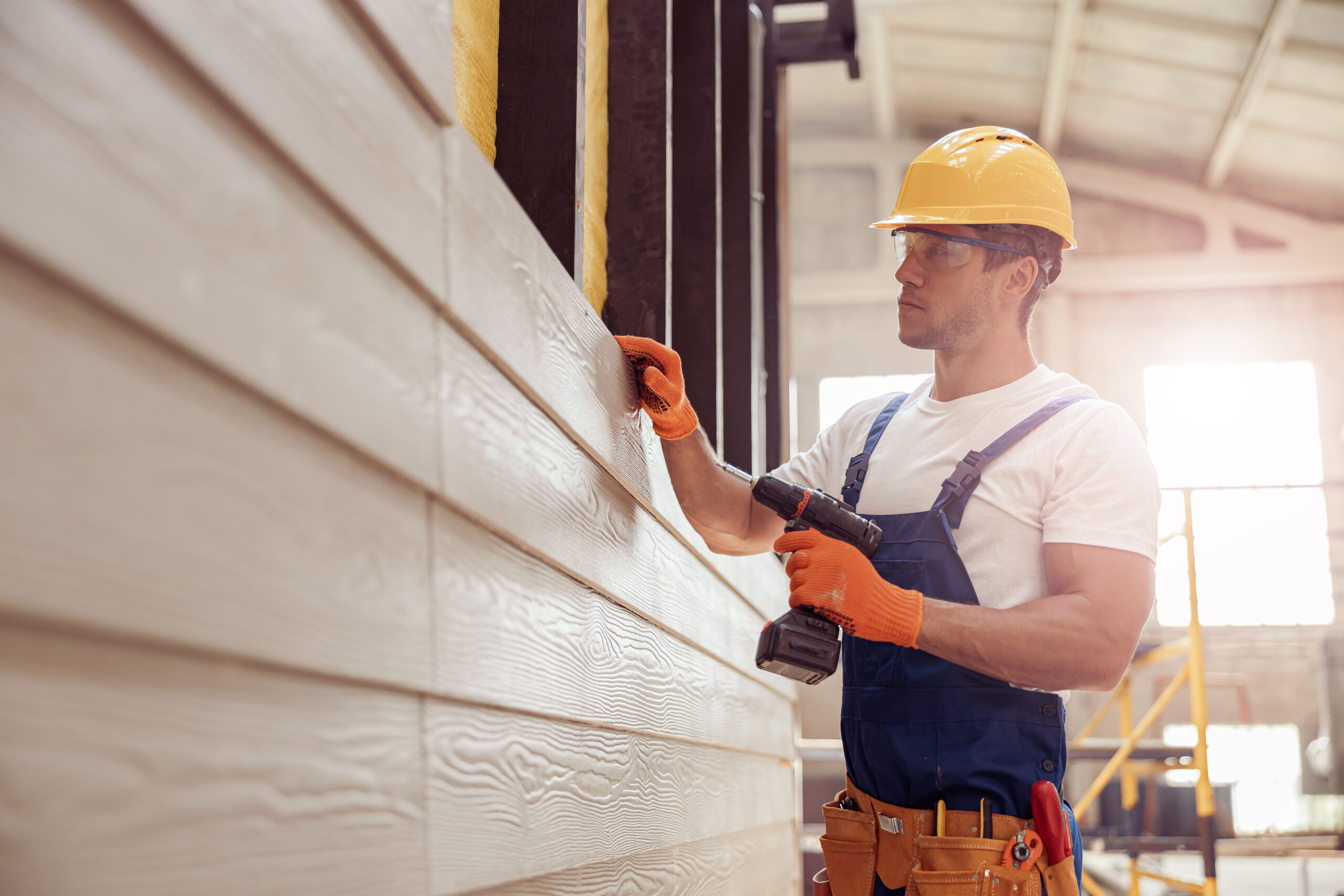 Male construction worker in safety helmet holding electric drill and checking wooden plank while building house